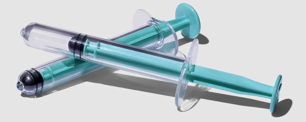 Two transparent syringes with teal accents positioned diagonally on a light gray background.