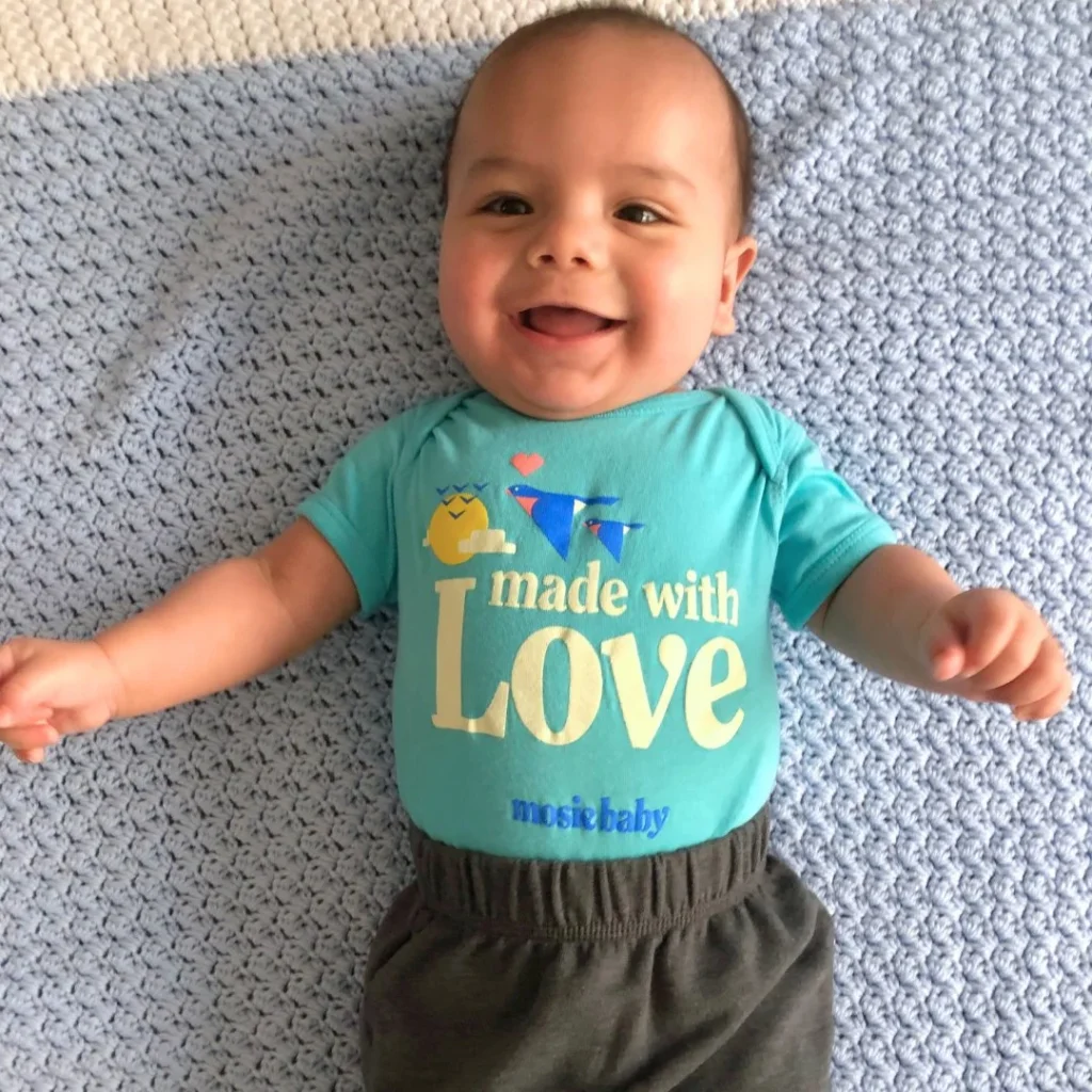 Smiling baby in a blue onesie with "made with Love" text, lying on a light blue crocheted blanket.