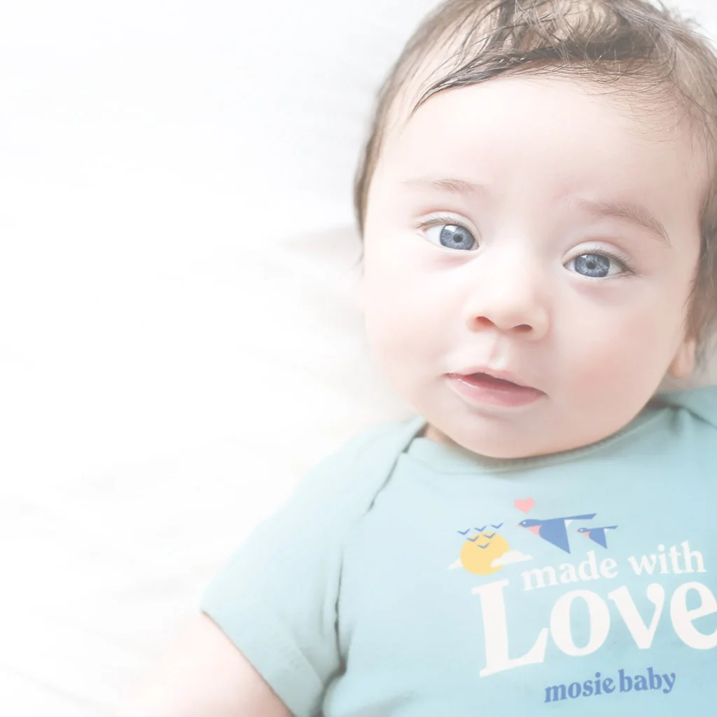 Baby with blue eyes wearing a light blue onesie that says "made with Love" on a soft white background.